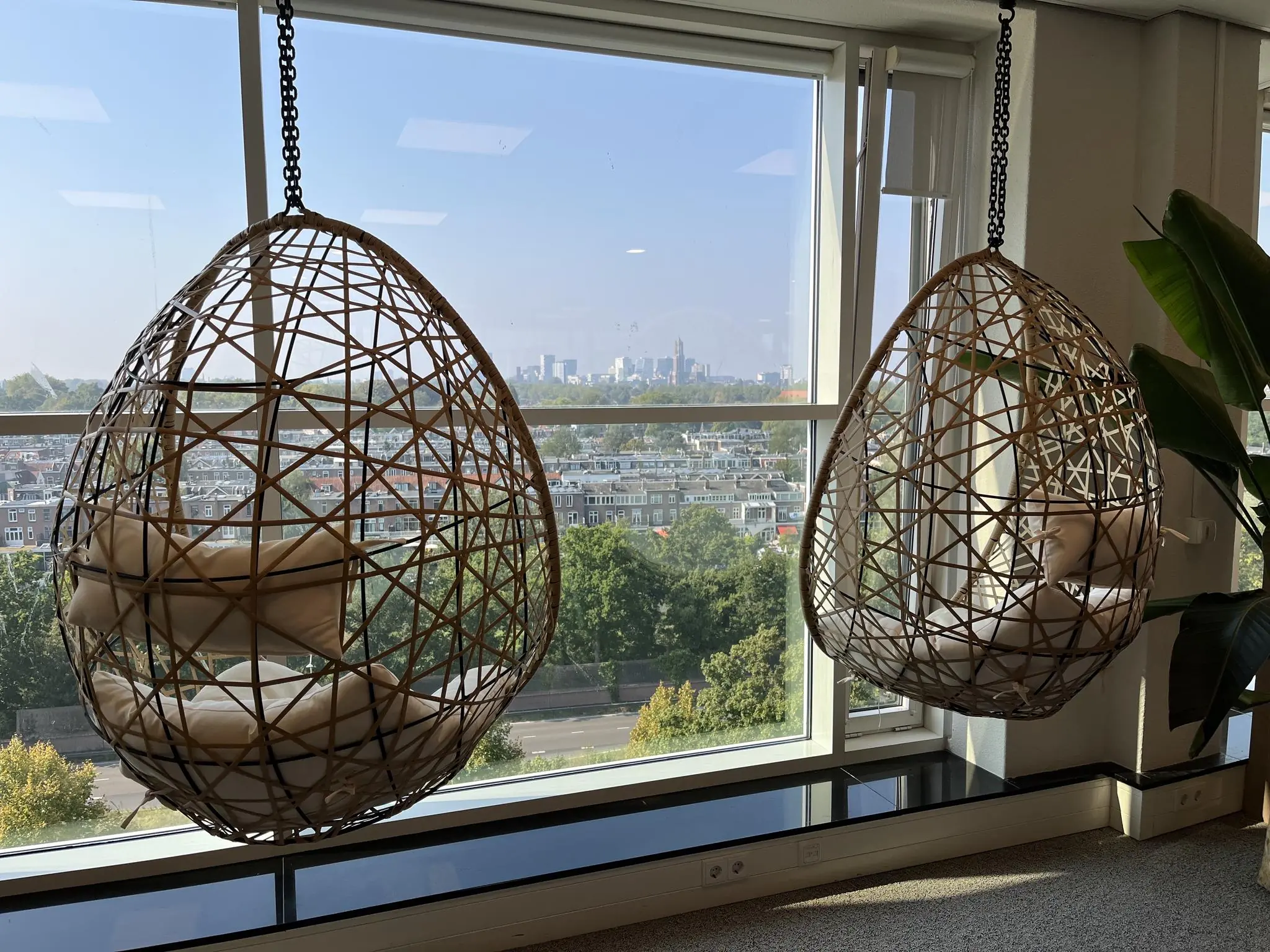 Twee hangstoelen van rotan met kussens hangen voor een groot raam met uitzicht op de stad vanaf Newtonlaan.