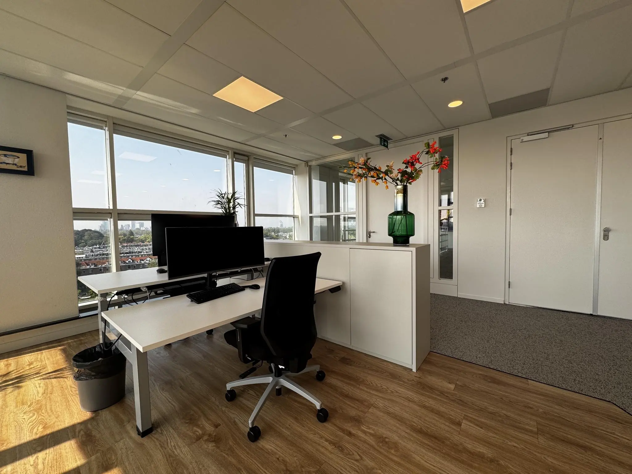 Modern kantoorinterieur aan de Newtonlaan met werkplek bij het raam, houten vloer en bloemenvaas op een kast.