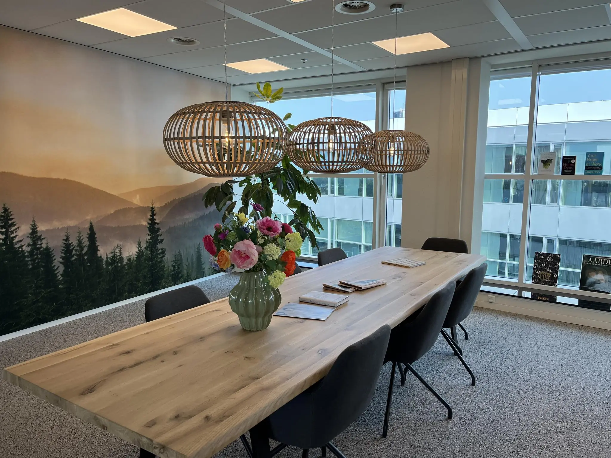 Vergaderruimte aan de Newtonlaan met een houten tafel, boeketten bloemen, natuurwanddecoratie en uitzicht op kantoorpanden.