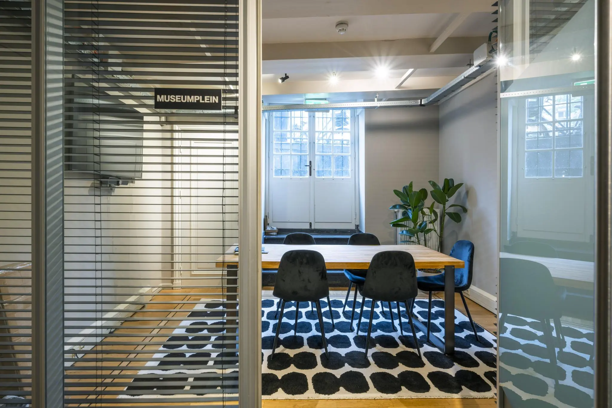 Vergaderruimte ‘Museumplein’ met houten tafel, zes stoelen, zwart-wit vloerkleed en een plant bij het raam.