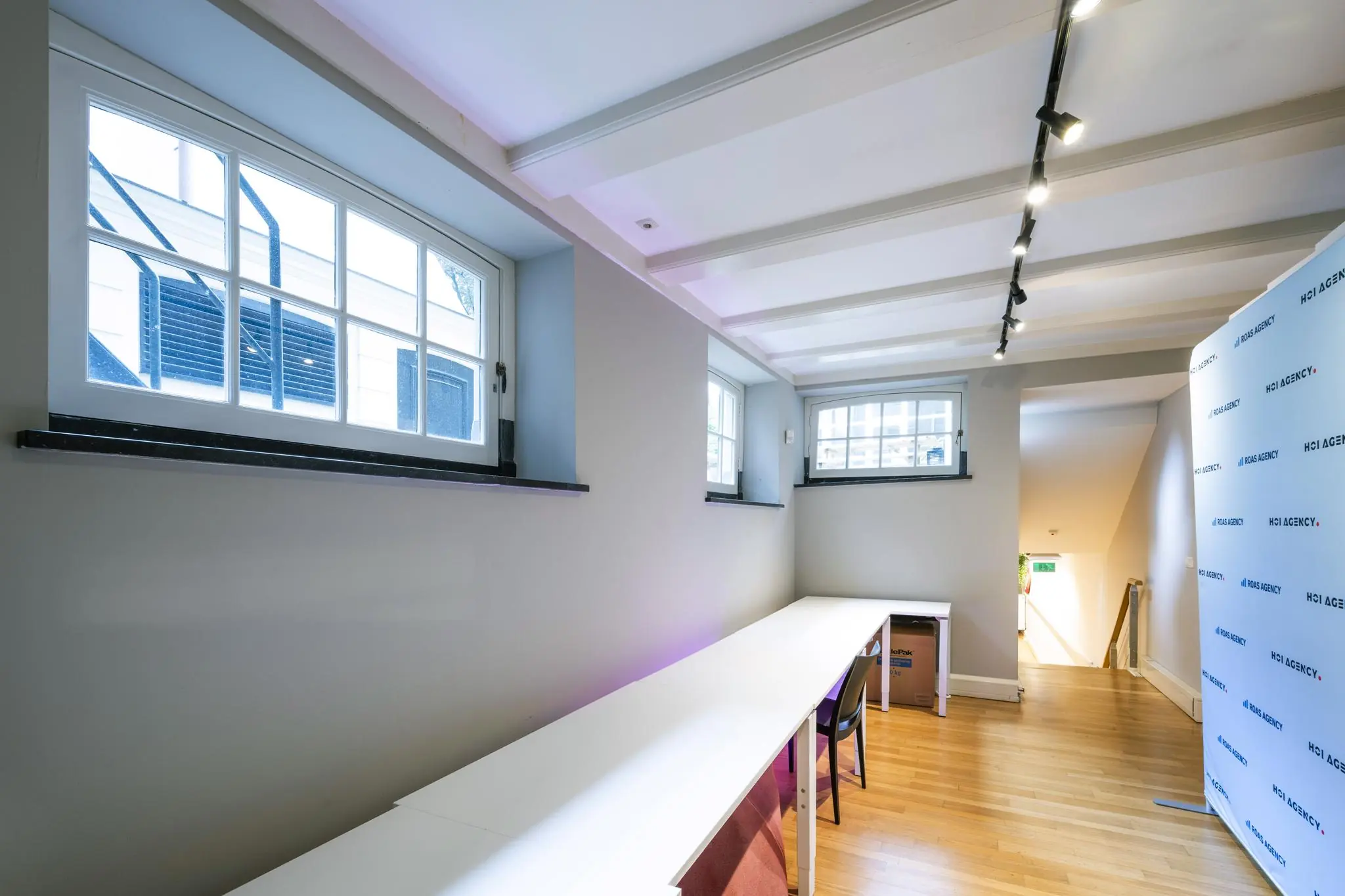 Licht souterrainkantoor aan de Herengracht met lange witte werktafels, houten vloer en meerdere ramen.