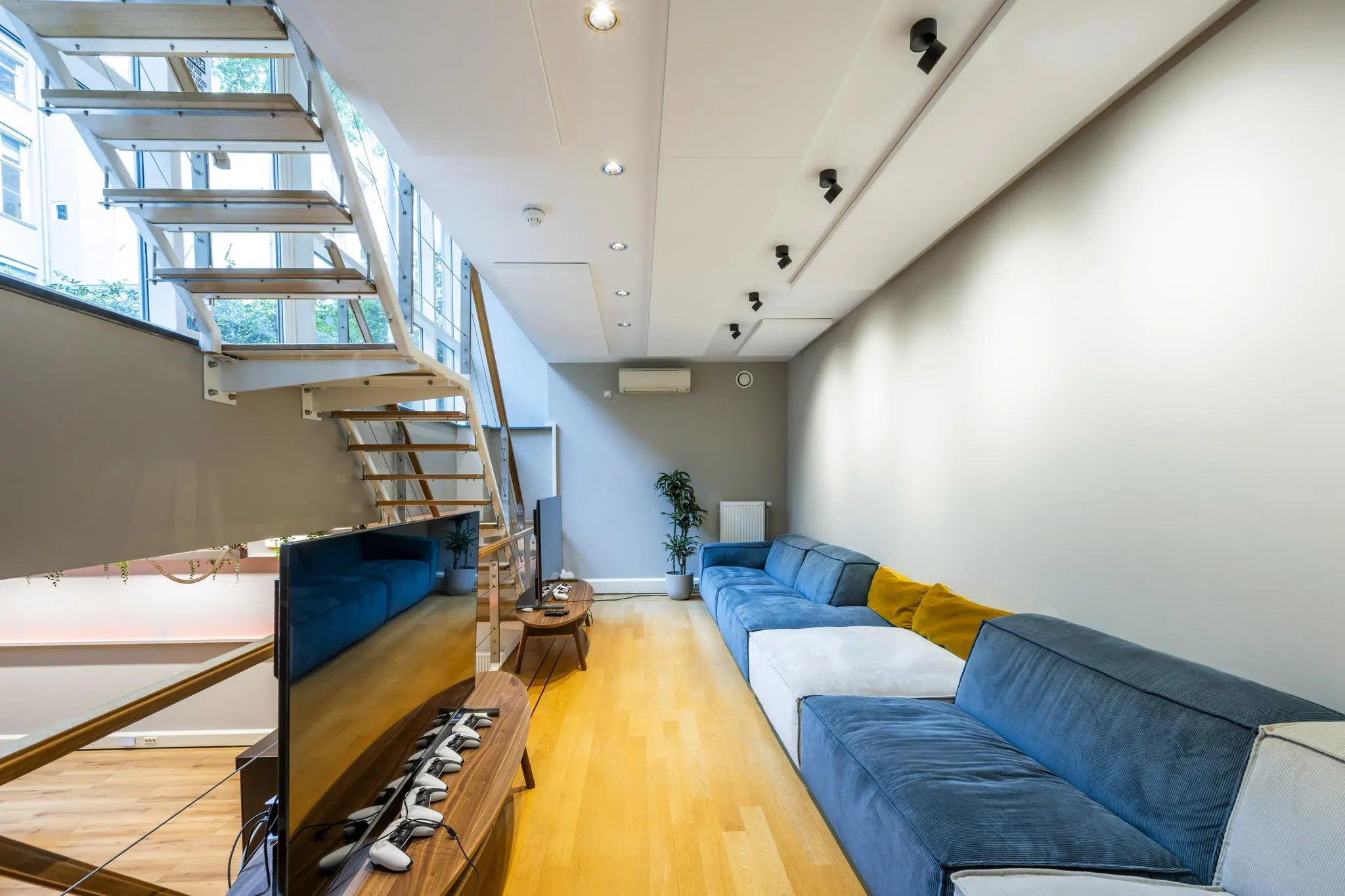 Moderne woonkamer aan de Herengracht met een grote blauwe bank, houten trap en een flatscreen-tv met meerdere gamecontrollers.