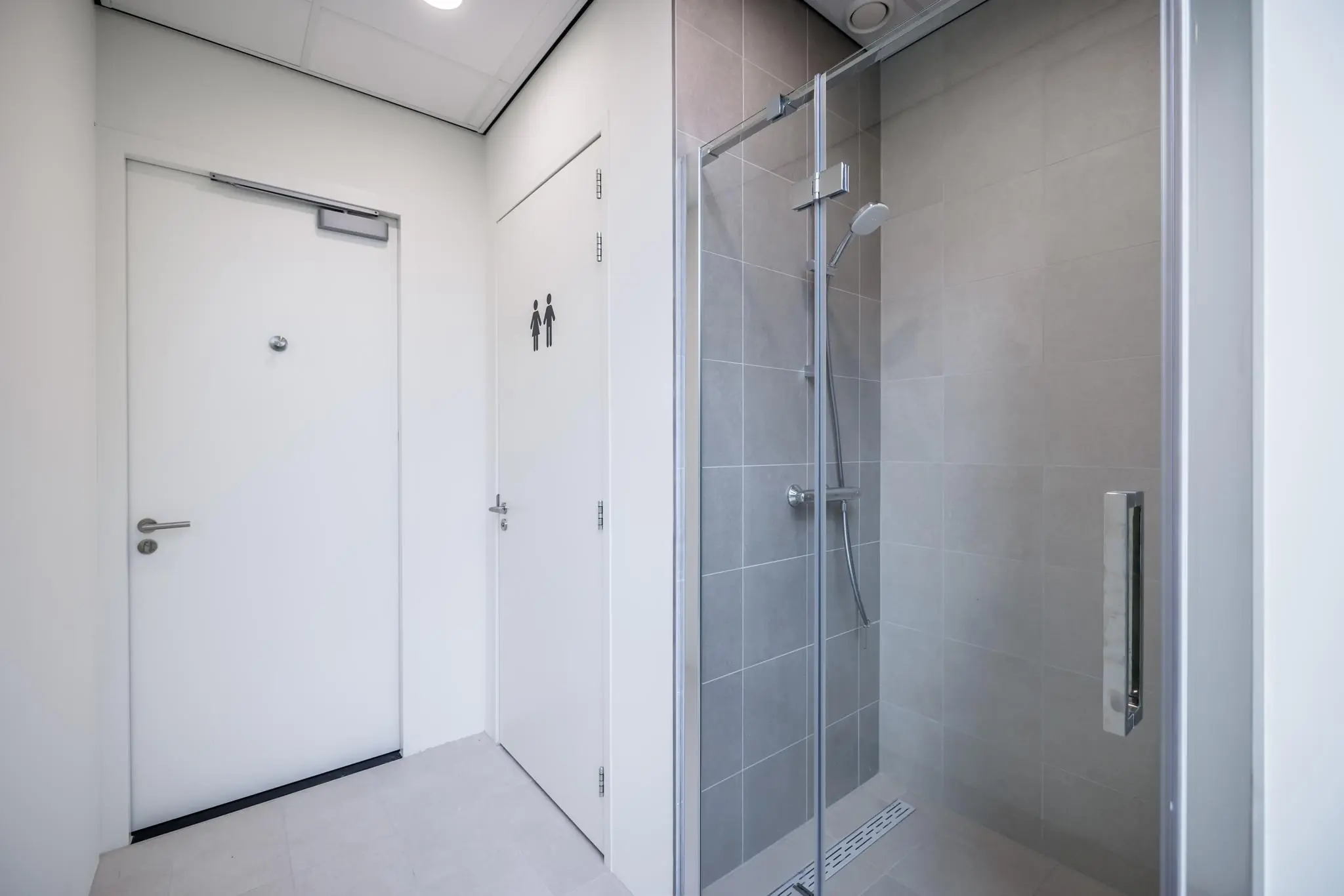 Moderne sanitaire ruimte met douche en twee gesloten deuren, waarvan één gemarkeerd als uniseks toilet.