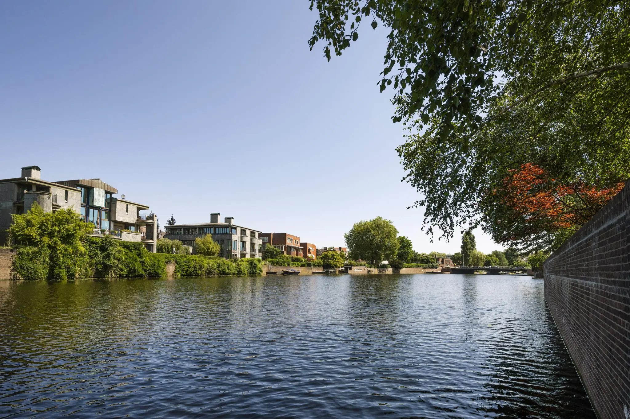 Moderne huizen aan het water langs de Apollolaan in Amsterdam, met een helderblauwe lucht en groene begroeiing.