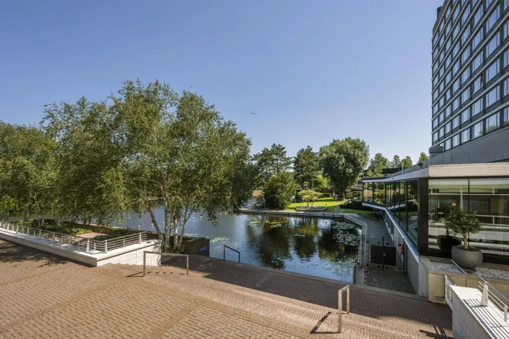 Modern gebouw aan de Apollolaan met uitzicht op een vijver en groene omgeving op een zonnige dag.