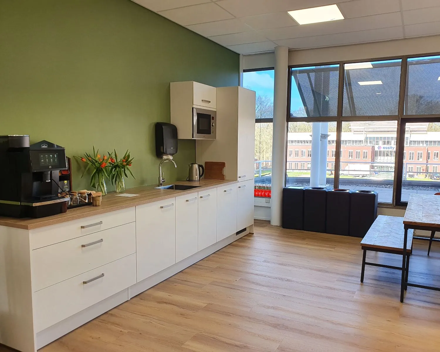 Moderne kantoorkeuken met koffiezetapparaat, magnetron en grote ramen met uitzicht op een kantoorgebouw.