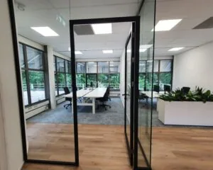 Moderne vergaderruimte met glazen wanden, bureaus en bureaustoelen, uitkijkend op een groene buitenomgeving.