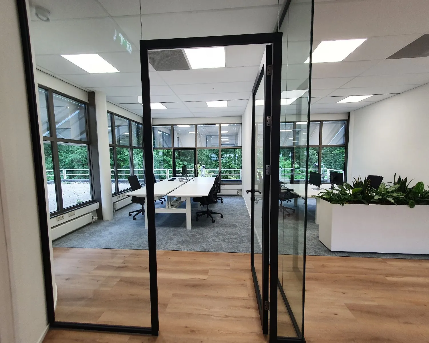 Moderne vergaderruimte met glazen wanden, bureaus en bureaustoelen, uitkijkend op een groene buitenomgeving.