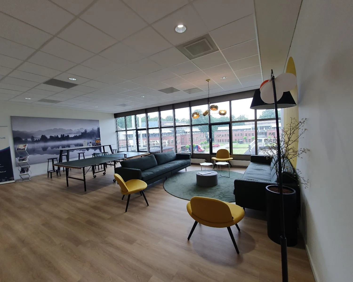 Moderne kantoorlounge met zithoeken, een pingpongtafel en grote ramen met uitzicht op een schoolachtig gebouw.
