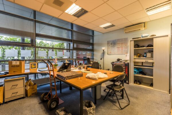 Kantoorruimte met grote ramen, een vergadertafel vol spullen en een open archiefkast, vermoedelijk in gebouw Veldzigt.