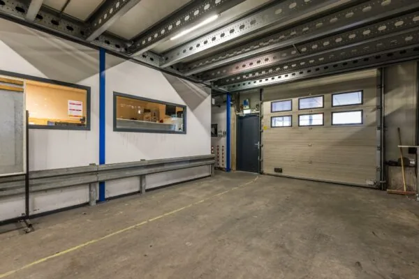 Interieur van een industriële loods met metalen plafondbalken, binnenramen en een gesloten rolpoort.