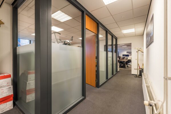 Kantoorhal met glazen kantoorwanden, openstaande houten deur en zicht op werkplekken in de verte.