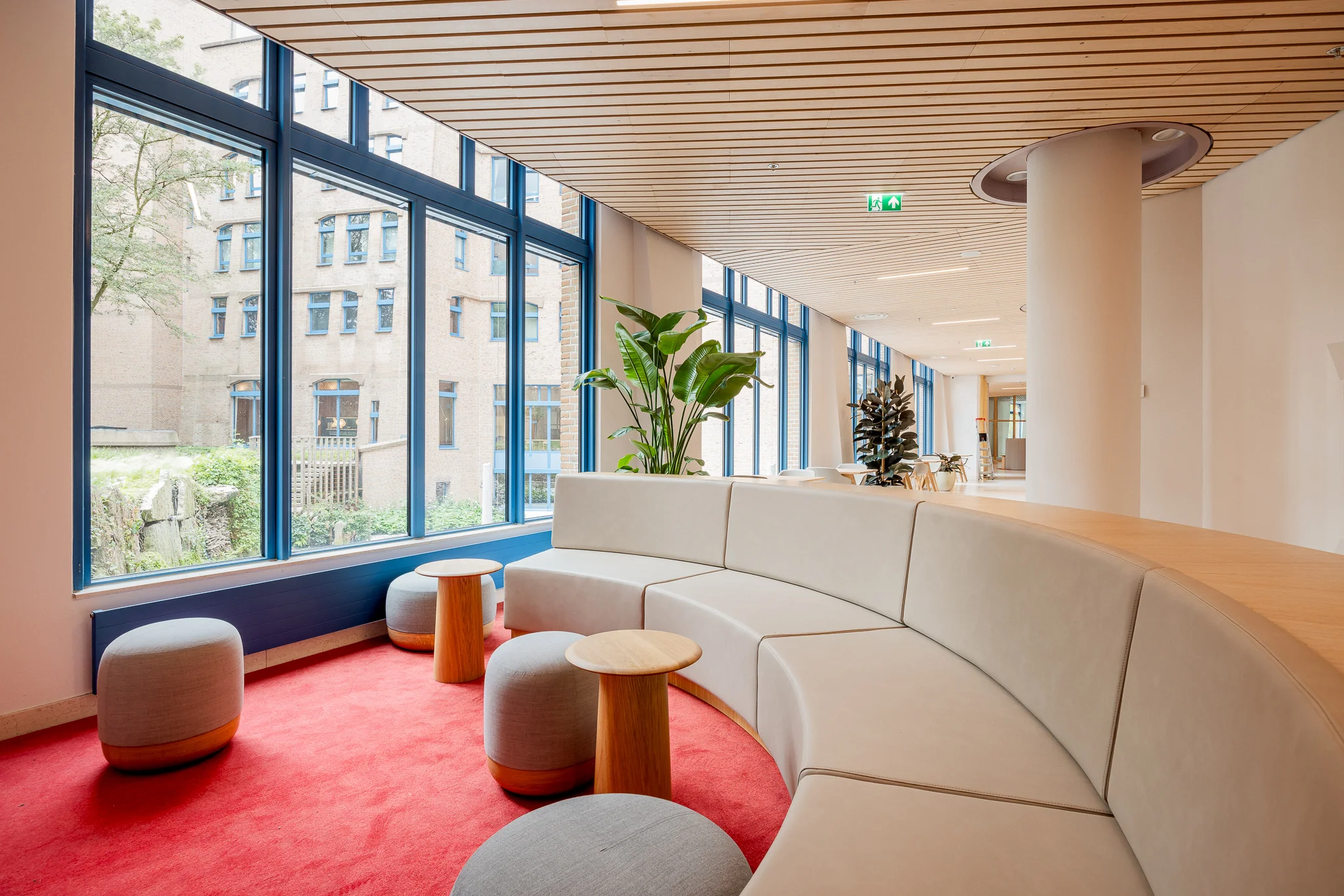 Moderne wachtruimte in het Zandkasteel met gebogen beige bank, rode vloerbedekking en grote ramen met uitzicht op de binnenplaats.