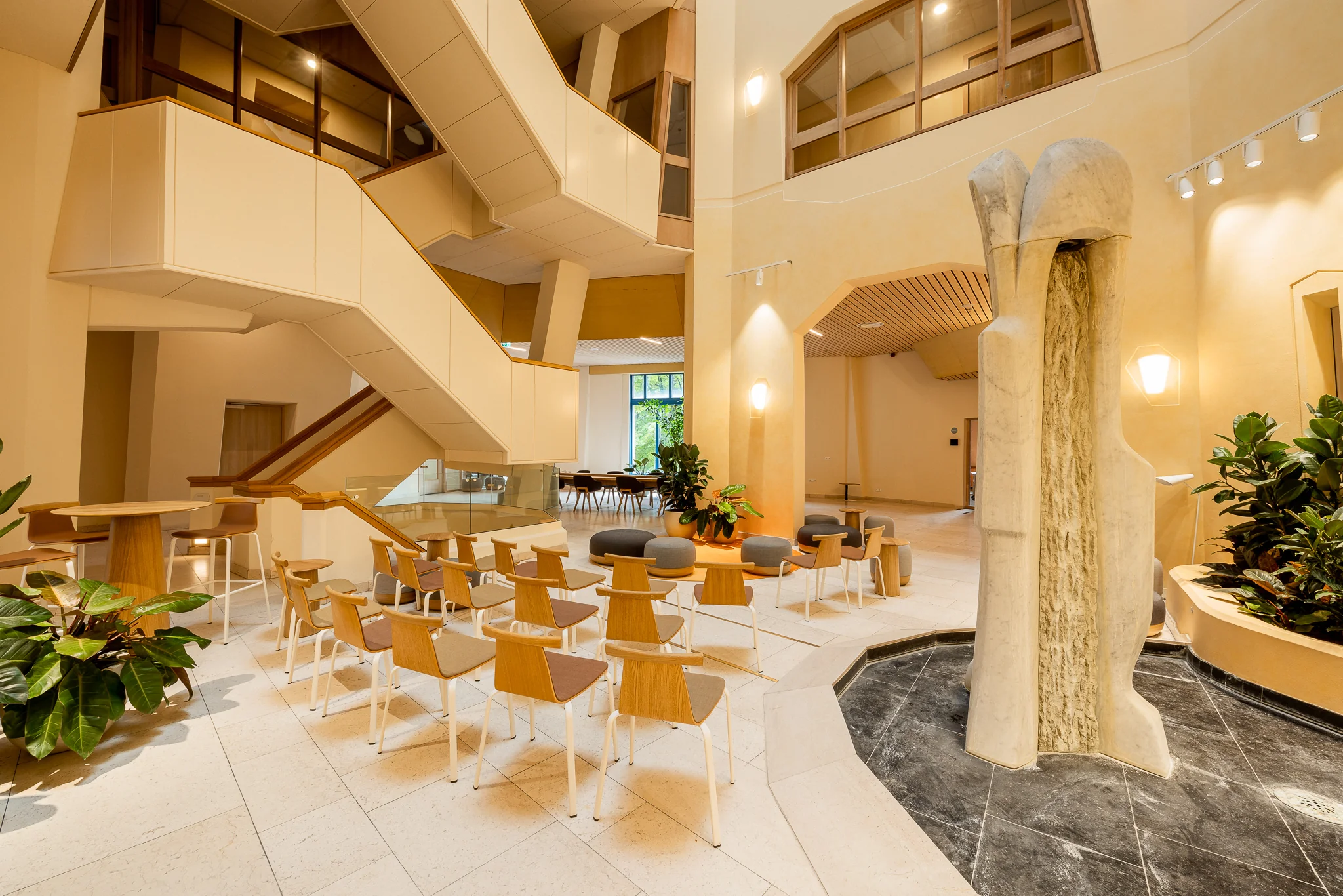 Lichte en moderne binnenruimte van Het Zandkasteel met houten stoelen, planten, sculpturale fontein en opvallend trappenhuis.
