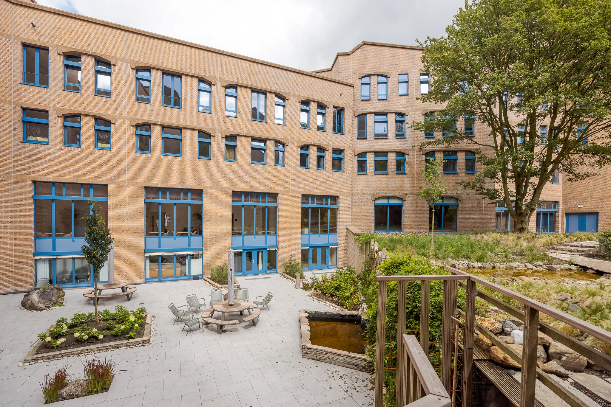 Binnenplaats van het Zandkasteel met zitgedeelte, plantenbakken en vijver, omringd door een bakstenen gebouw met blauwe kozijnen.