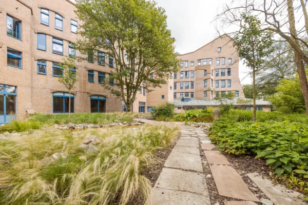 Binnenplaats van het Zandkasteel in Amsterdam met een kronkelend tuinpad, omringd door groen en het karakteristieke bakstenen gebouw met blauwe kozijnen.