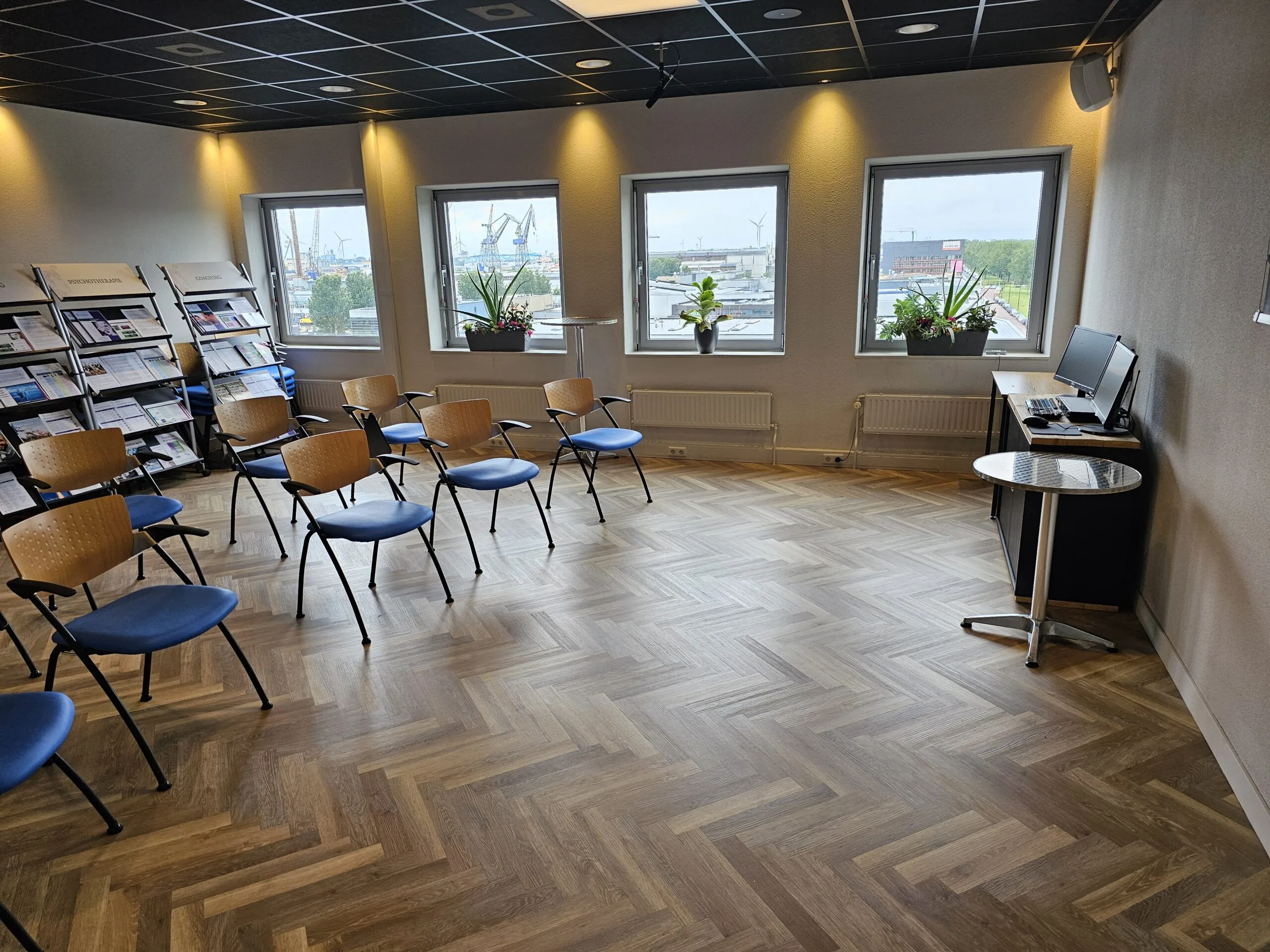 Vergaderruimte met opgestelde stoelen, een leestafel met folders en een computerhoek, met uitzicht op een industrieterrein door de ramen.