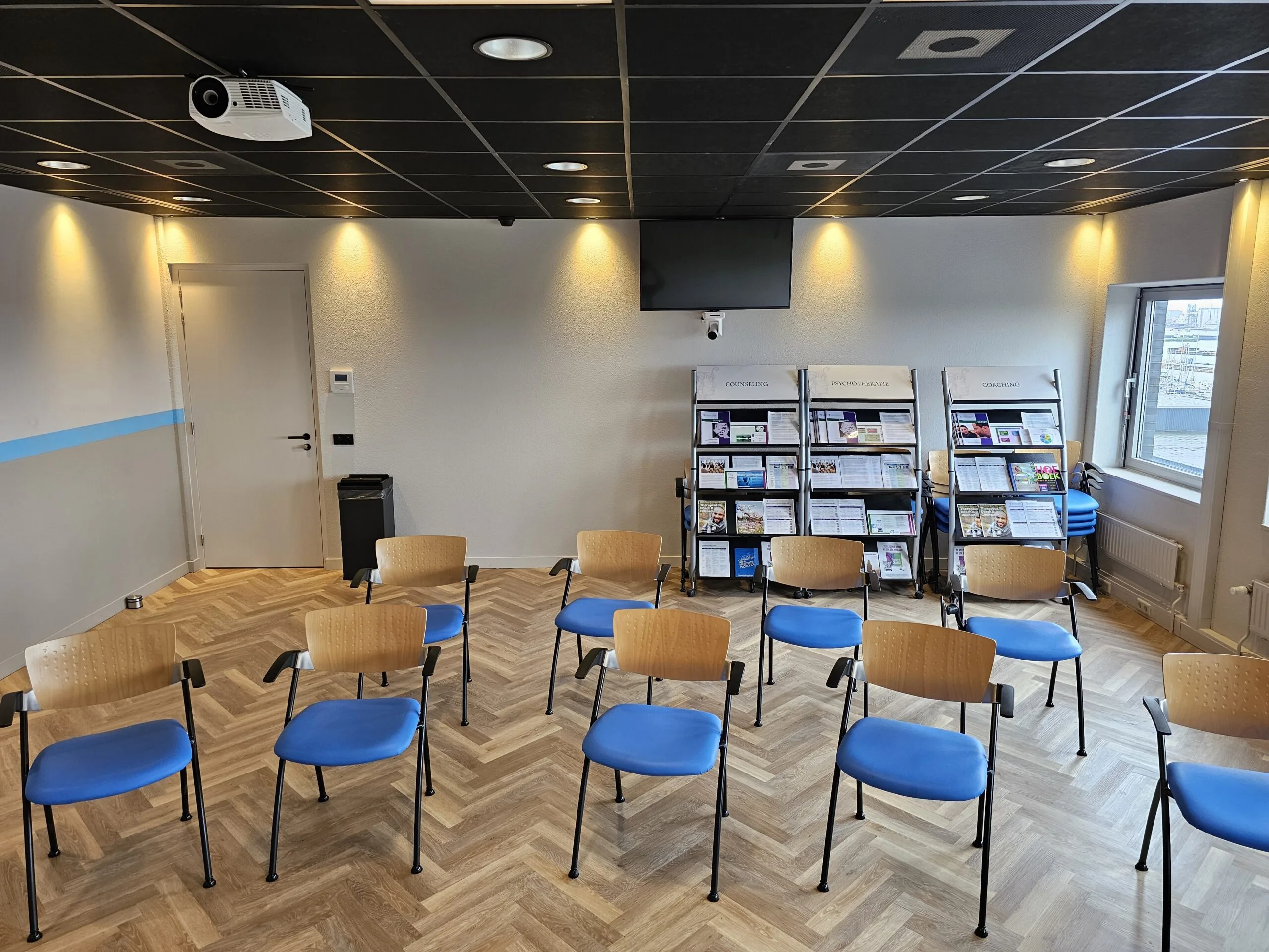 Wachtruimte of presentatieruimte met opgestelde stoelen, informatierekken en een scherm aan de muur.