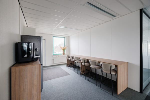 Een kantoorruimte met een lange houten tafel, hoge stoelen en een koffiezetapparaat naast een raam.
