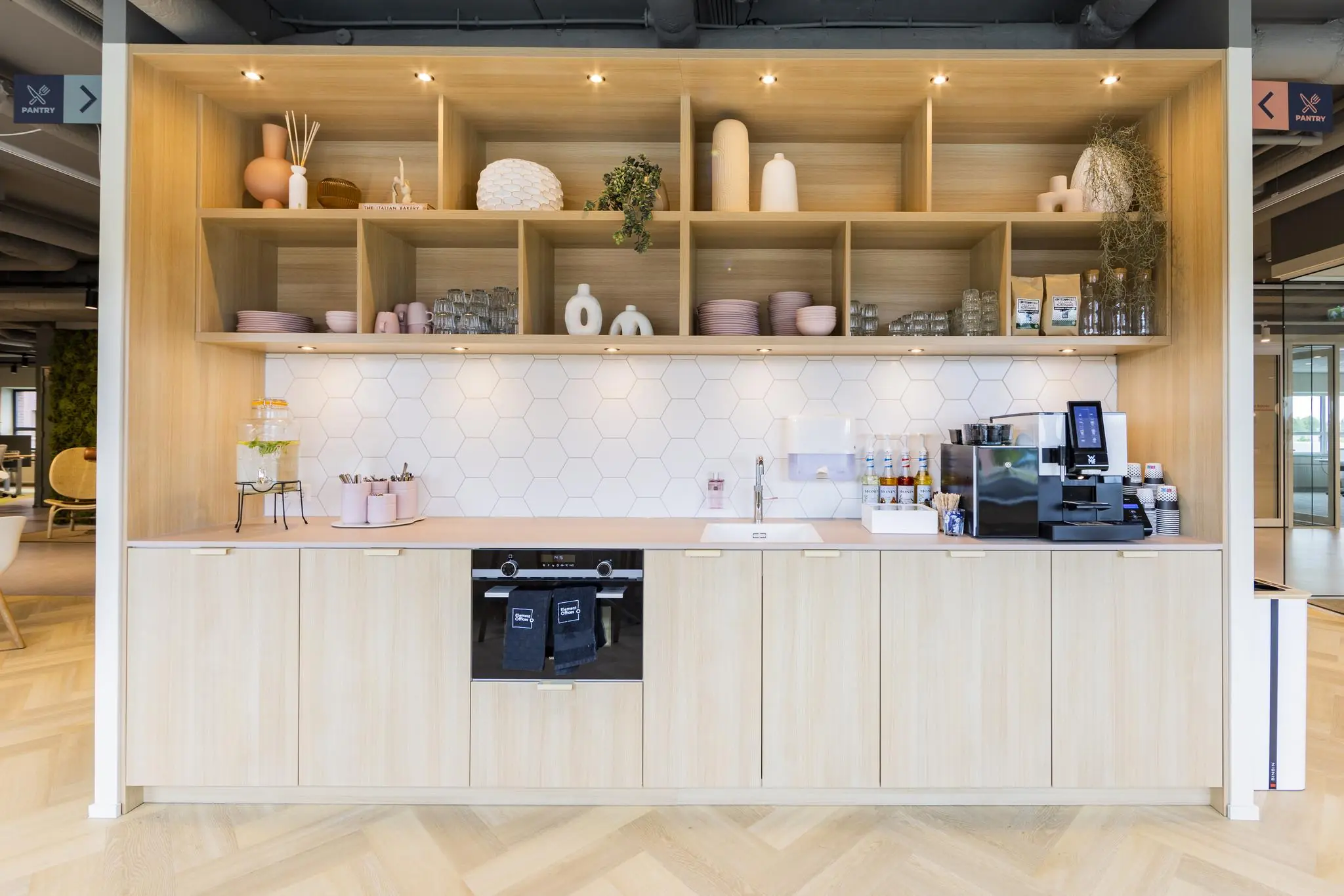 Moderne pantry met lichte houten kasten, open schappen, een koffiezetapparaat en een witte zeshoekige tegelwand.