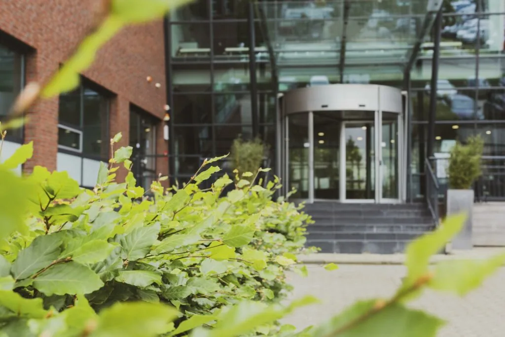 Vooraanzicht van het kantoorpand Rijnzathe met een glazen entree en groene haag op de voorgrond.