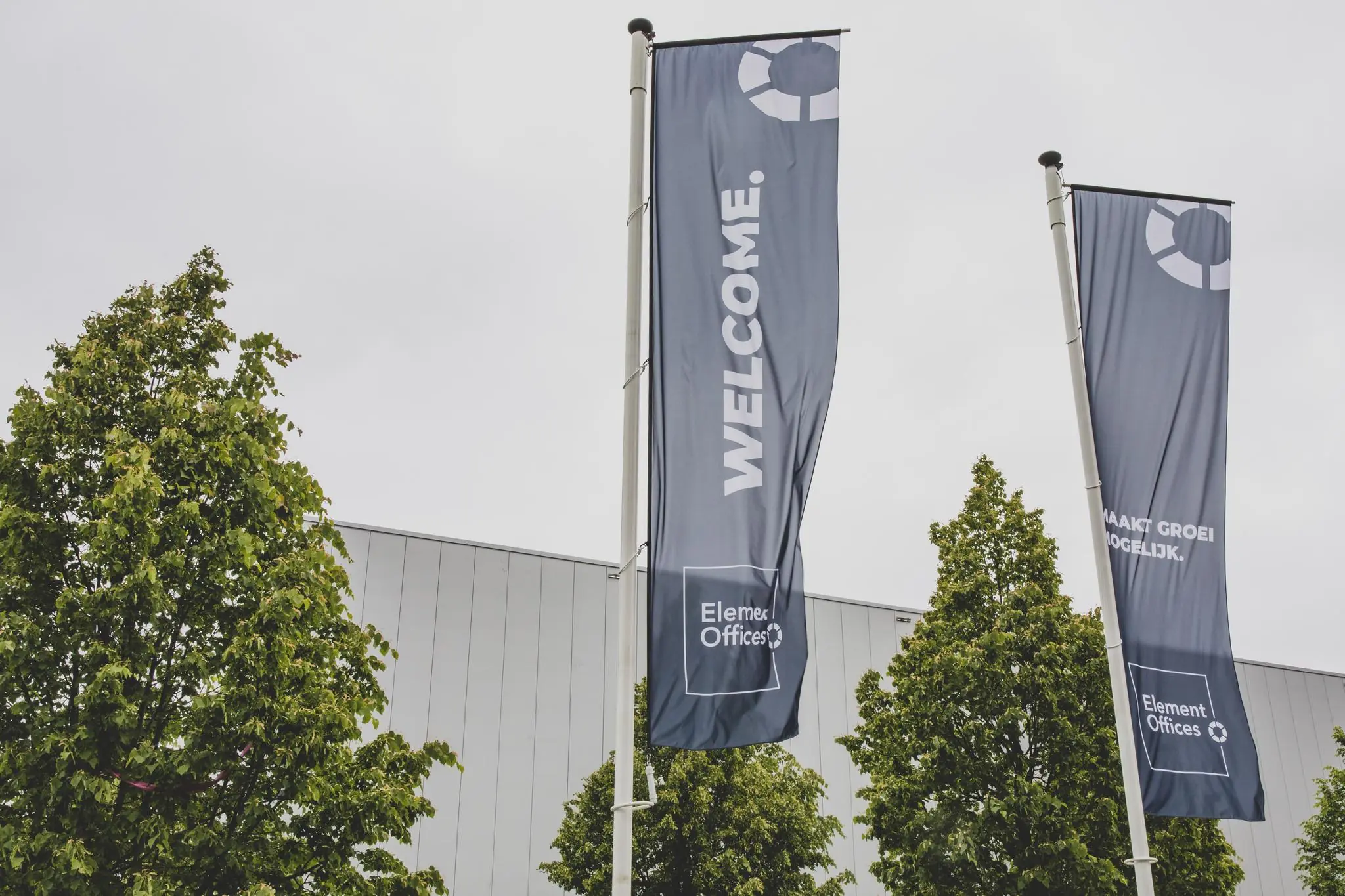 Vlaggen van Element Offices met de tekst "WELCOME" en "Maakt groei mogelijk" wapperen voor een modern kantoorgebouw in Rijnzathe.