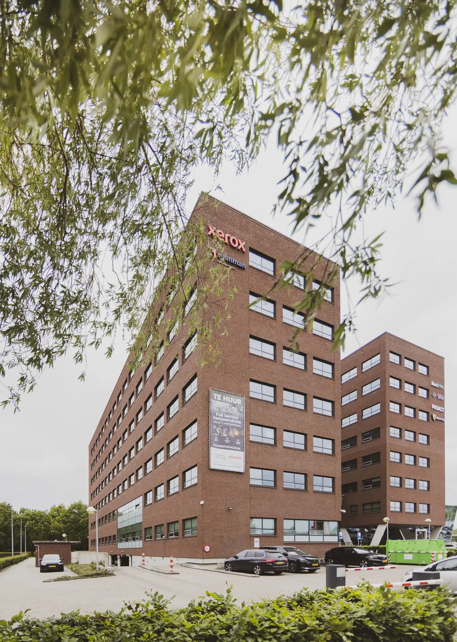 Kantoorgebouw Rijnzathe met onder meer de logo’s van Xerox, Hageman en Proact, gefotografeerd vanaf de parkeerplaats met bomen op de voorgrond.