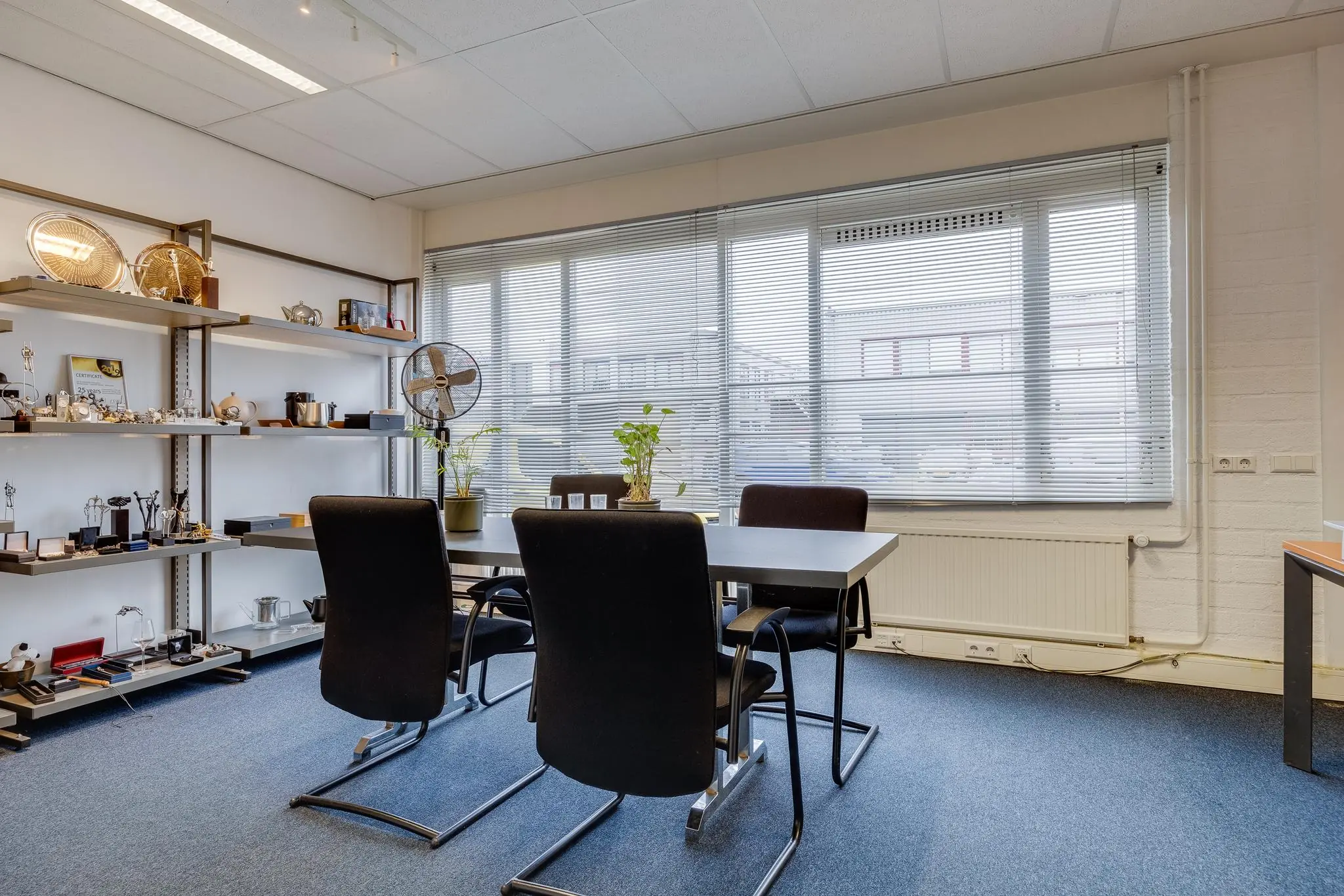 Vergaderruimte aan de Savannahweg met vier stoelen, een tafel, grote ramen met jaloezieën en een wandrek vol trofeeën en decoraties.