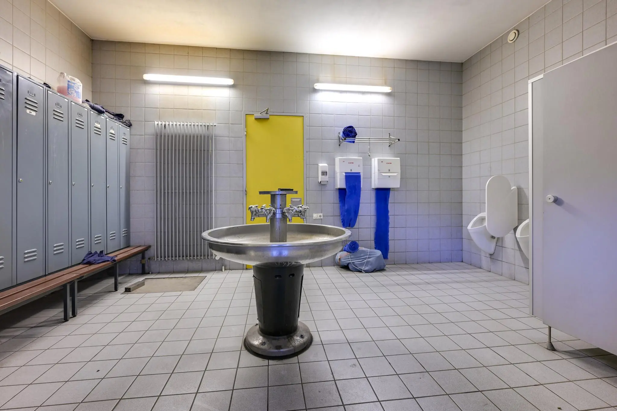 Kleedkamer met grijze lockers, centrale ronde wasbak, urinoirs en gele deur in Tennesseedreef gebouw.