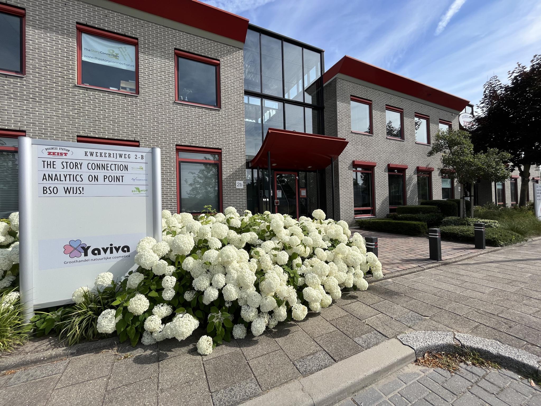 Bedrijventerrein aan de Kwekerijweg 2-B in Zeist met een kantoorgebouw en een bord met bedrijfsnamen omringd door bloeiende hortensia’s.