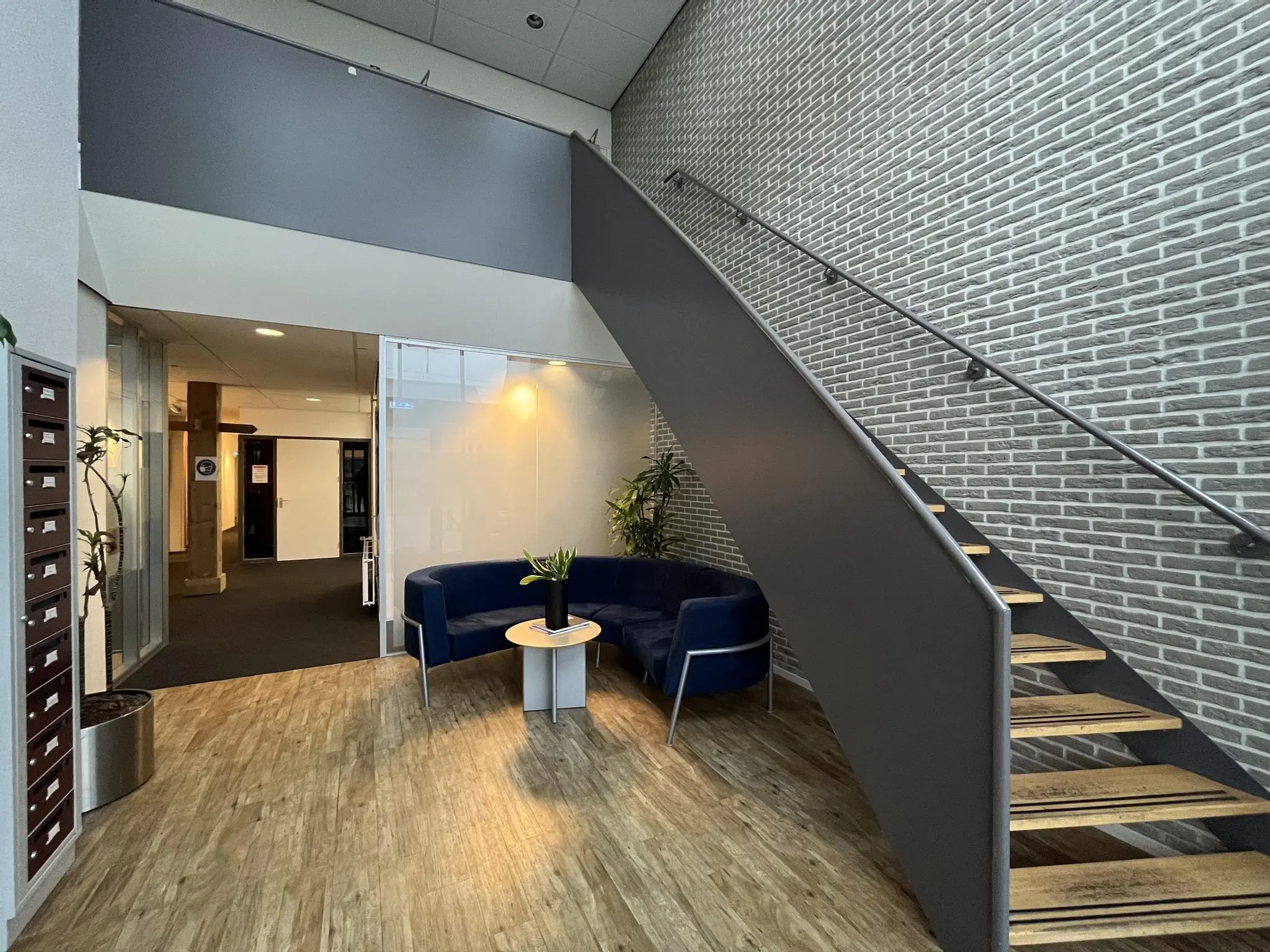 Moderne entreehal aan de Kwekerijweg met houten vloer, blauwe zithoek en een zwarte trap langs een grijze bakstenen muur.