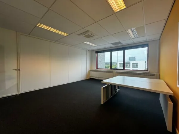 Leeg kantoor met wit bureau, grote ramen en uitzicht op omliggende gebouwen aan de Kwekerijweg.