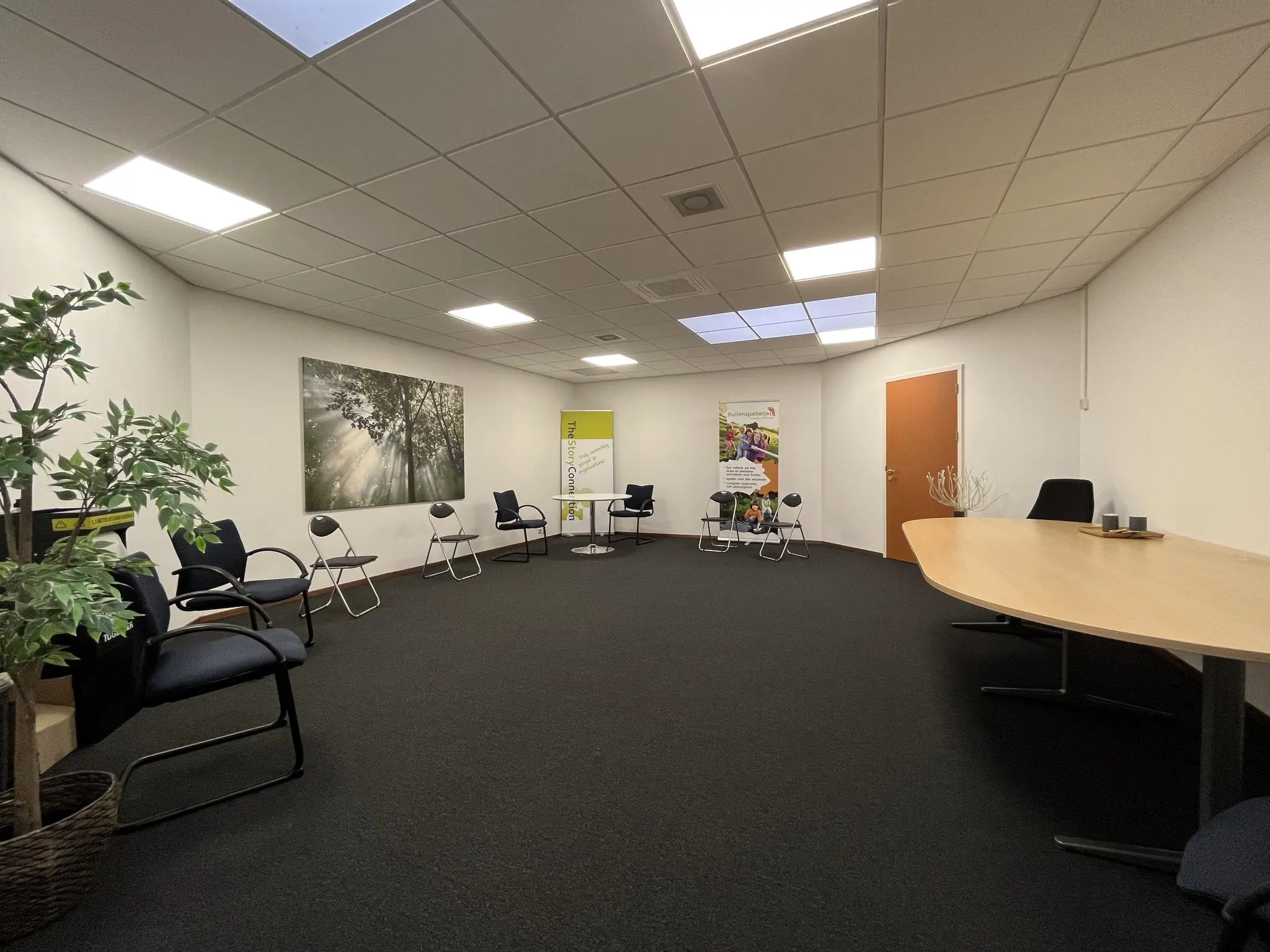 Vergaderruimte aan de Kwekerijweg met stoelen in een kring, een ovale tafel, planten en promotiebanners tegen een witte muur.