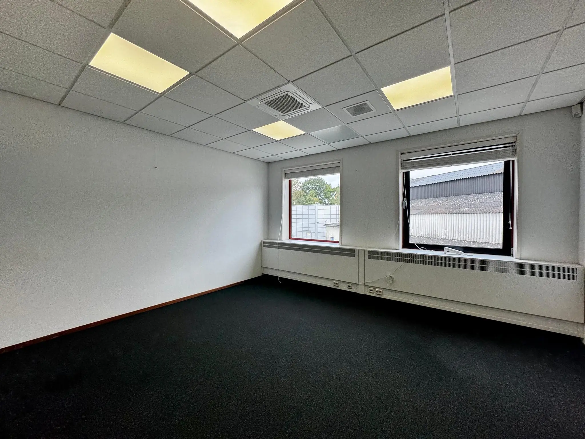 Leeg kantoorruimte aan de Kwekerijweg met witte muren, systeemplafond en twee ramen met deels gesloten jaloezieën.