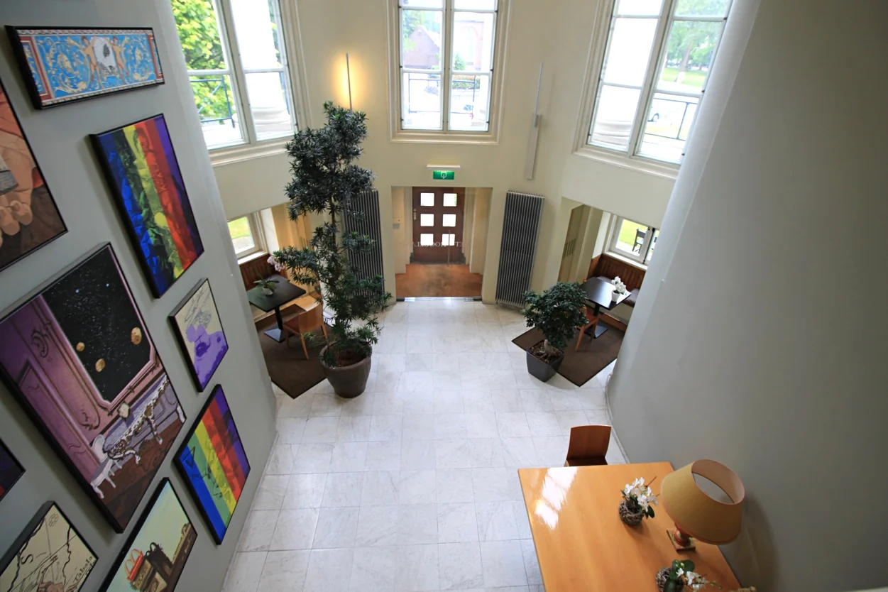 Inkomhal van Museumlaan met grote ramen, kunstwerken aan de muur en moderne inrichting met planten en zitjes.