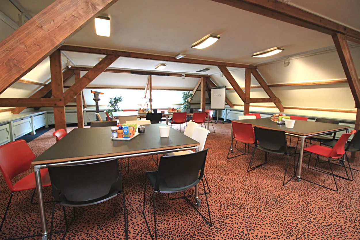 Vergaderruimte op Museumlaan met houten balken, kleurrijke stoelen en tafels met koffievoorzieningen.