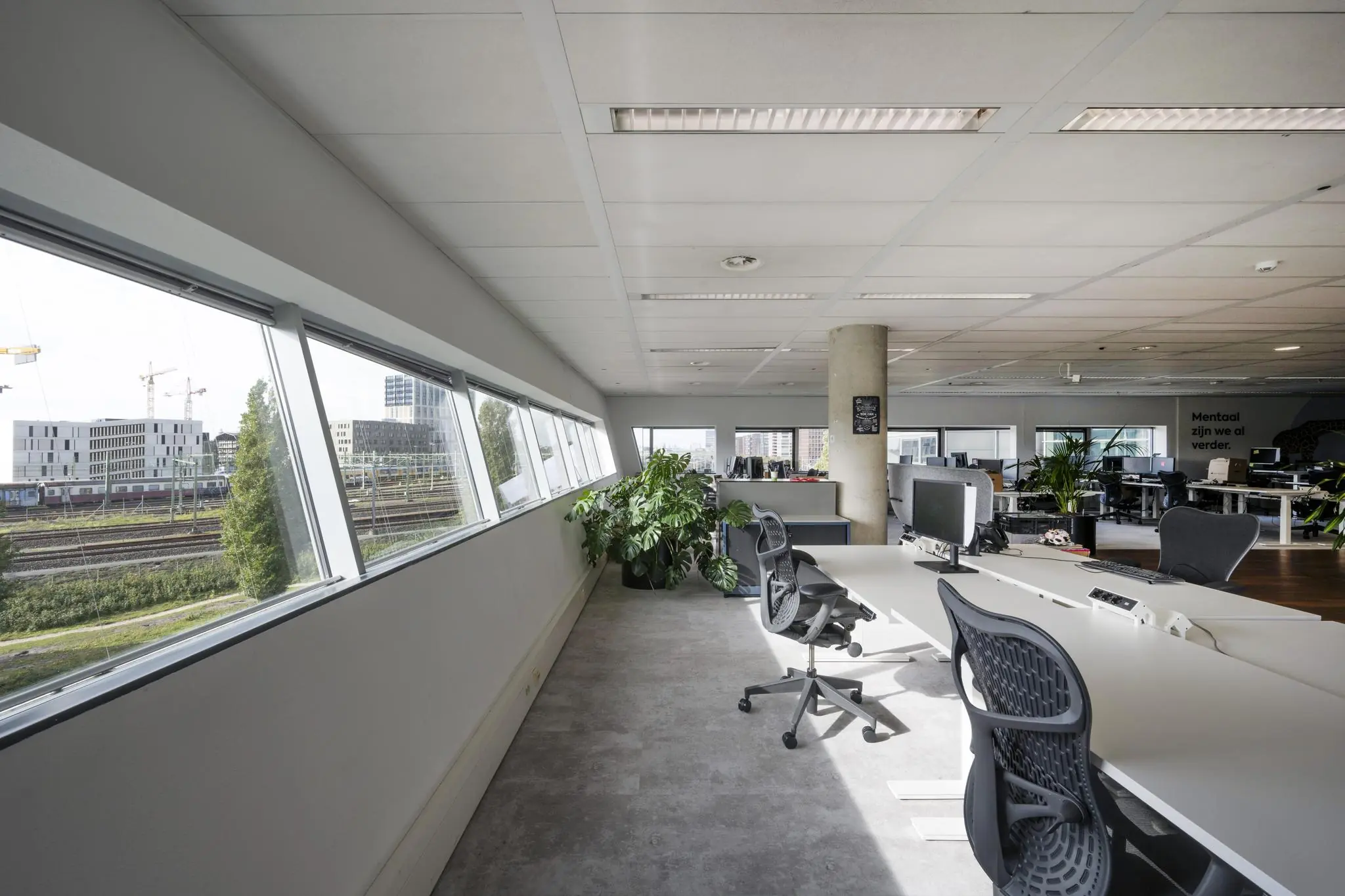 Modern kantoorinterieur in Rietlandpark met werkplekken, bureaustoelen en uitzicht op spoorlijnen en gebouwen.