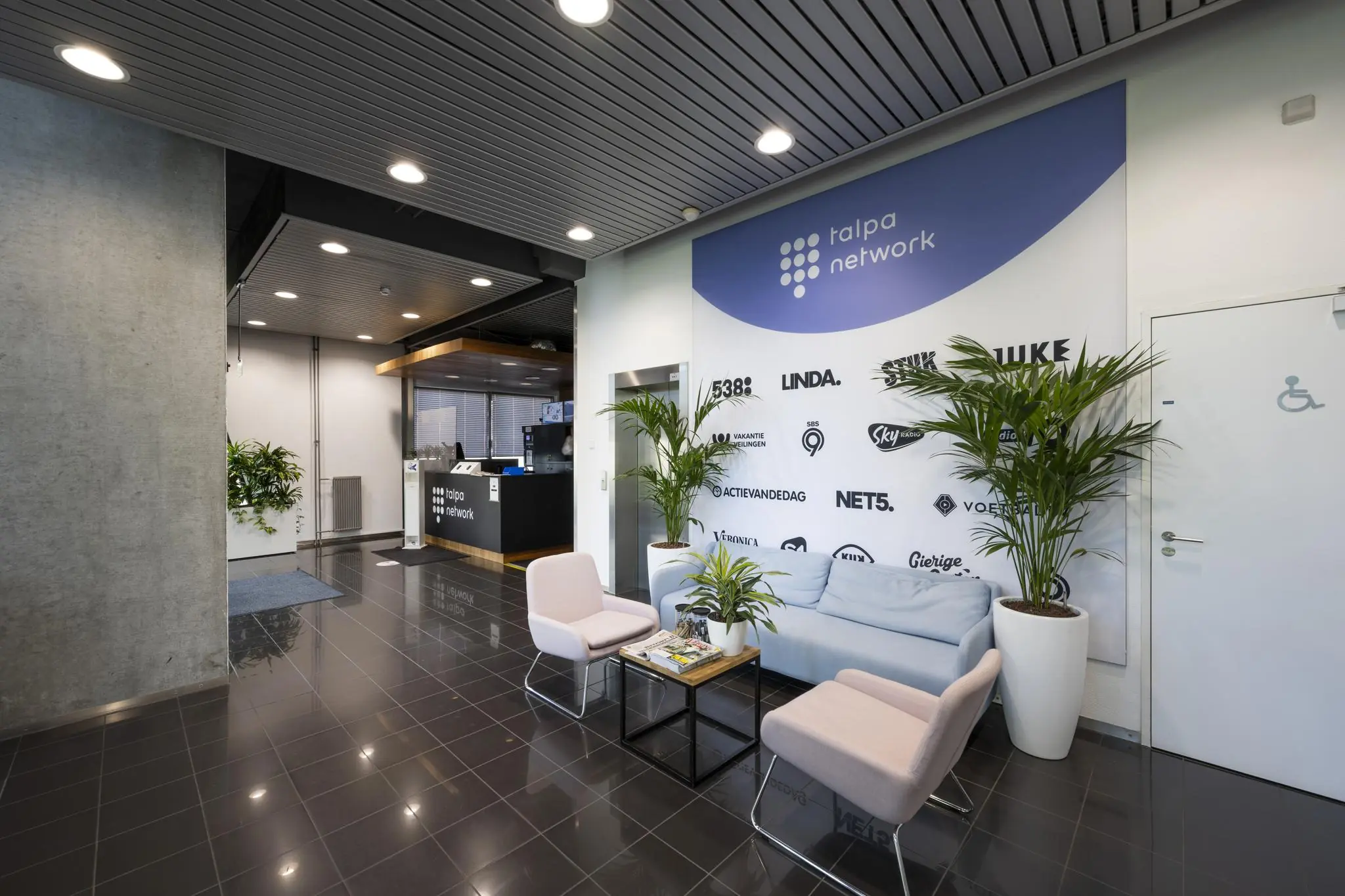 Moderne wachtruimte bij Talpa Network met zitbank, stoelen, planten en een wand met logo’s van mediakanalen.