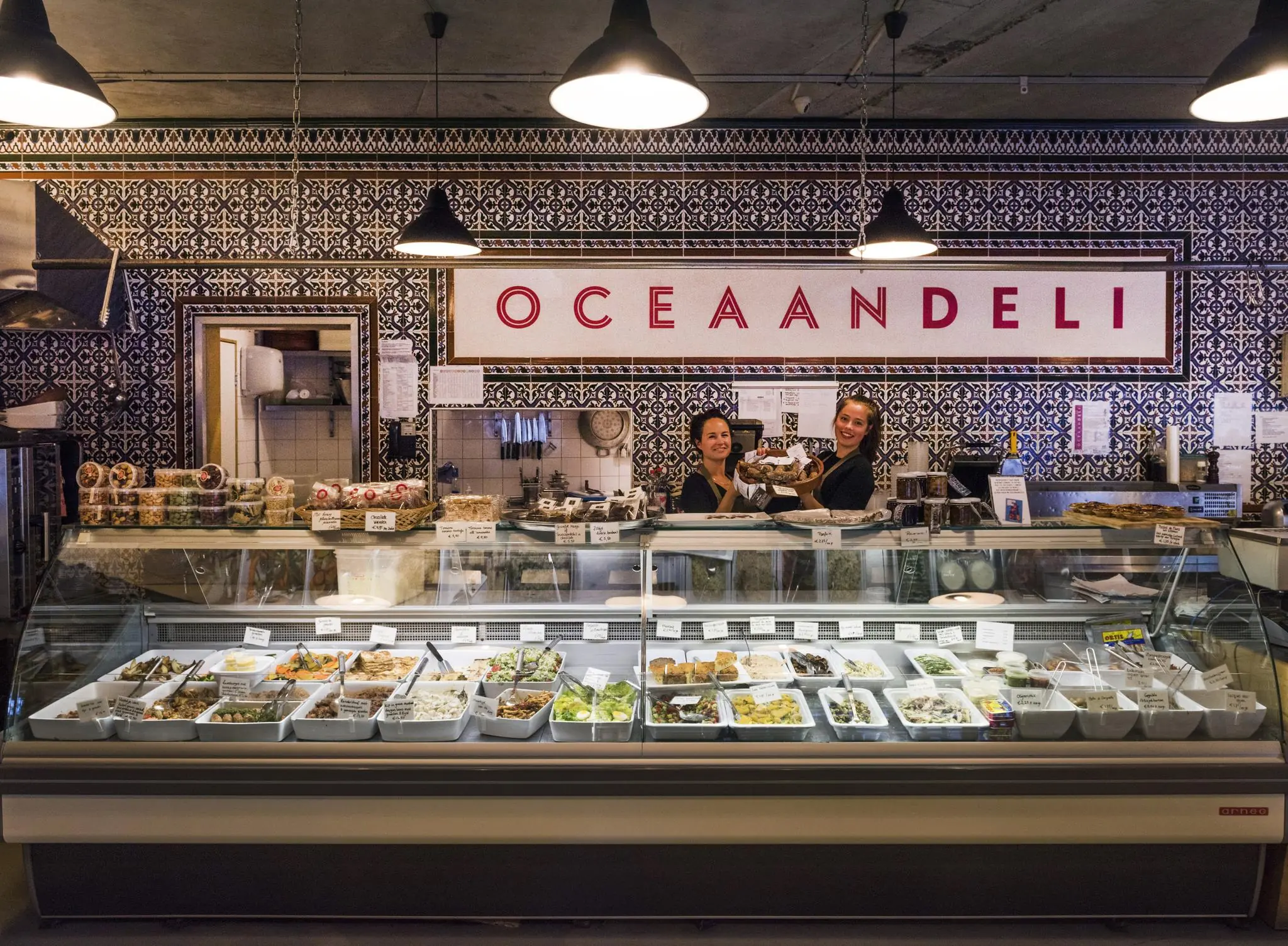 Twee medewerkers achter de toonbank van Oceaan Deli in Rietlandpark, met een vitrine vol mediterrane gerechten.