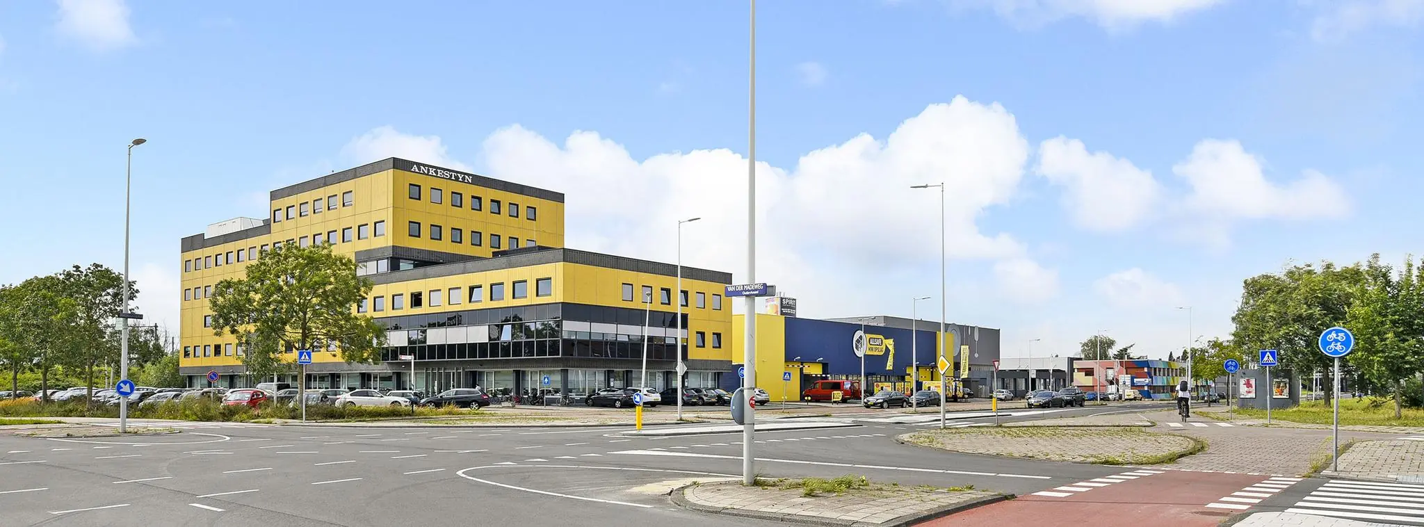 Kantoorgebouw AnkeStyn en winkelpand aan de Joan Muyskenweg in Amsterdam met rotonde en fietspad op de voorgrond.