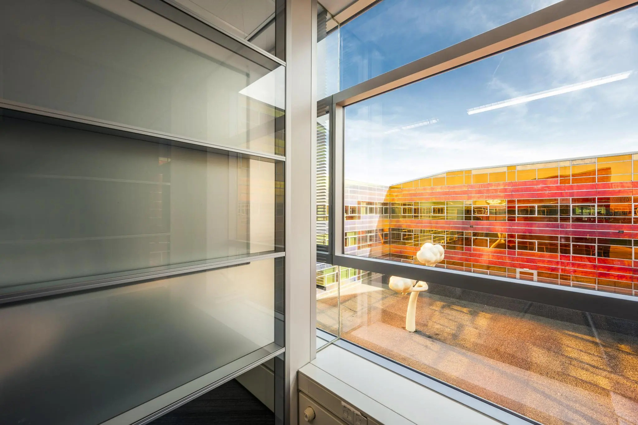 Zicht vanuit een kantoorruimte aan de W. Dreesweg op een modern gebouw met gekleurde glazen gevels en kunstobjecten op het dak.