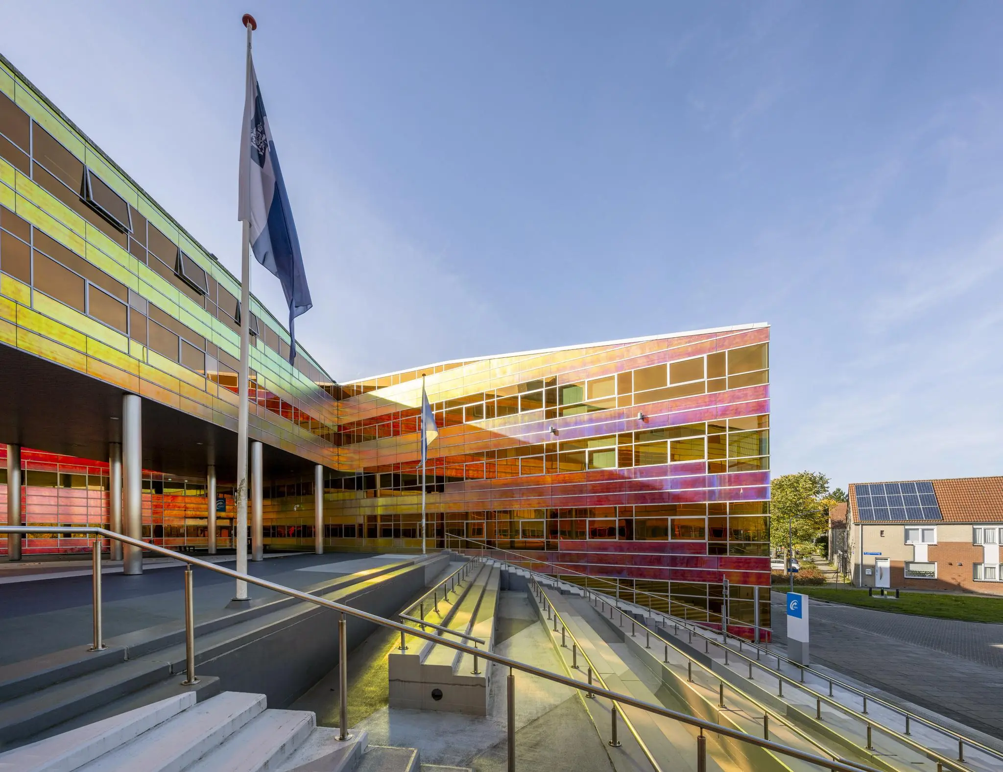 Modern gebouw met kleurrijke, reflecterende gevel aan de W. Dreesweg, met trap en vlaggenmast op de voorgrond.
