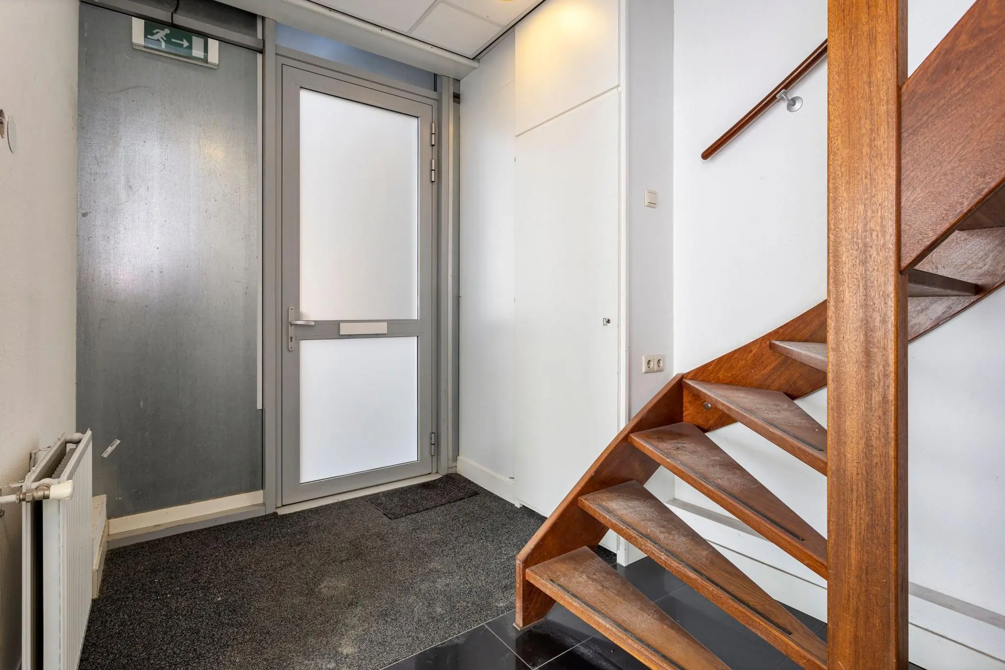 Ingangshal met grijze voordeur, tapijtvloer en houten trap in een woning aan de Tijnmuiden.