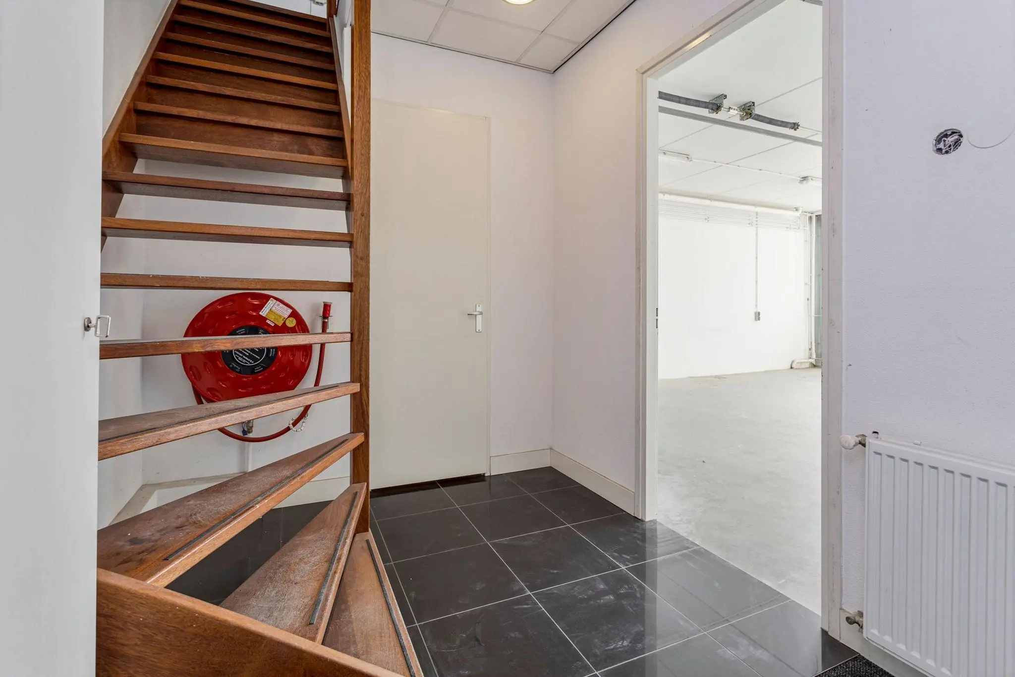 Ingang met houten trap, brandslanghaspel en deur naar een lichte, industriële ruimte in een pand aan de Tijnmuiden.