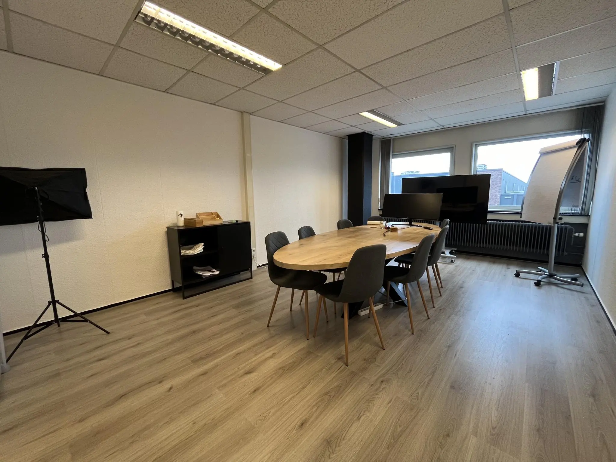 Vergaderruimte aan de Hollantlaan met ovale houten tafel, acht stoelen, televisieschermen en een flip-over bij het raam.