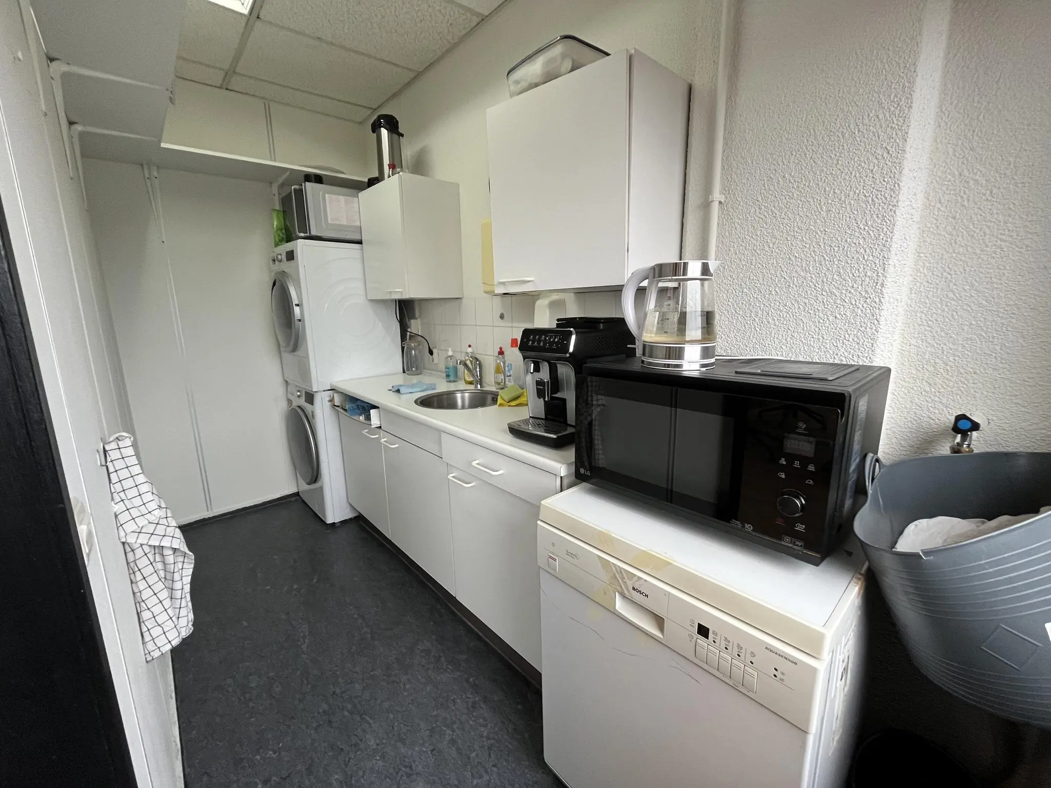 Kleine keukenruimte aan de Hollantlaan met wit keukenblok, stapelbare wasmachine en droger, magnetron, koffiezetapparaat en waterkoker.