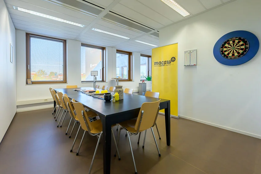 Vergaderruimte met lange tafel, acht stoelen, dartbord aan de muur en een gele banner met het logo van Macau.