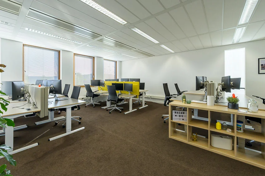 Modern kantoor met meerdere bureaus, computers en ergonomische bureaustoelen in een lichte ruimte met grote ramen.