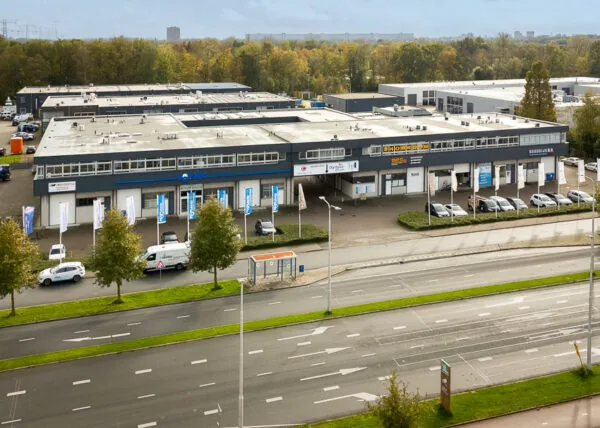 Bedrijfsverzamelgebouw aan de Hoofdweg met meerdere auto’s, vlaggen en een bushalte langs de weg.