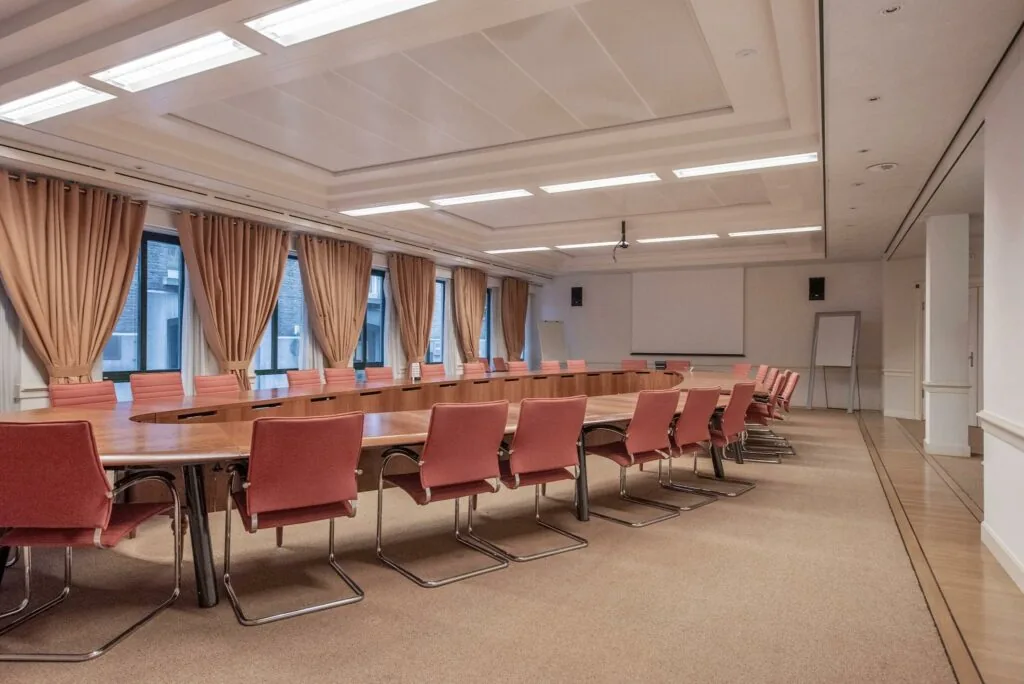 Ruime vergaderzaal aan de Doelenstraat met lange ovale tafel, roze stoelen en grote ramen met beige gordijnen.